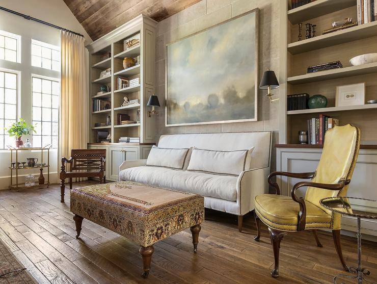 A large landscape art piece hangs over a white nailhead sofa placed facing a kilim bench and paired with a yellow leather French accent chair. The sofa is flanked by light gray built-in shelves mounted over light gray cabinets.