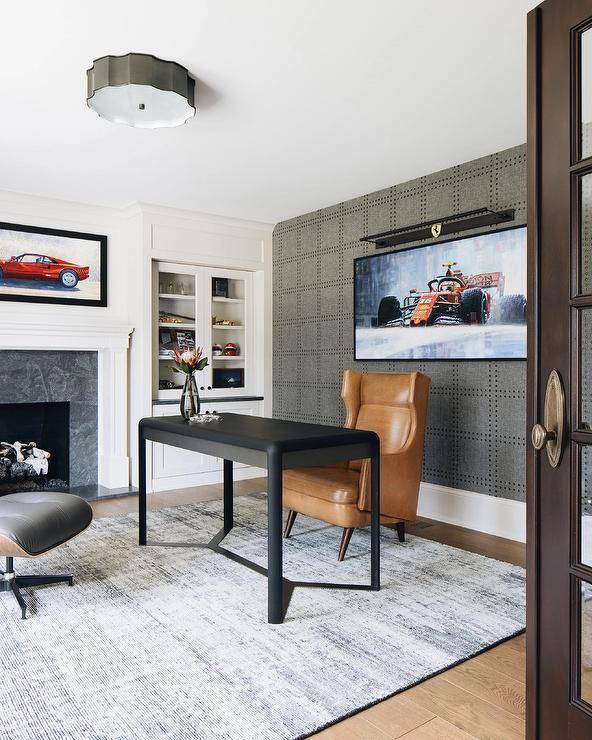 Masculine home office features an accent wall covered in Phillip Jeffries Rivet wallpaper and finished with a Samsung Frame TV. A caramel brown leather wingback task chair sits on a gray overdyed rug at a matte black desk. Glass front cabinets are built-in to the side of the desk and over white cabinets.
