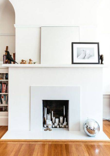 a neutral minimal faux fireplace with lots of various candles inside and very laconic and minimal decor on the mantel