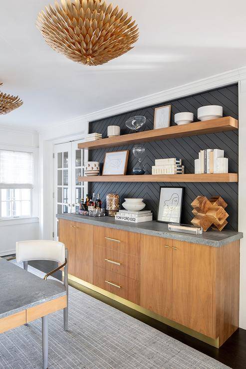 Brown veneer floating shelves are stacked against a black chevron plank backsplash over a gray marble countertop accenting brown veneer office cabinets.