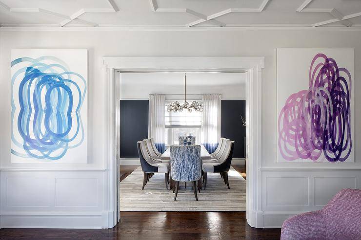 Blue and purple abstract art are hung in a contemporary living room over wainscoting and on either side of a doorway.