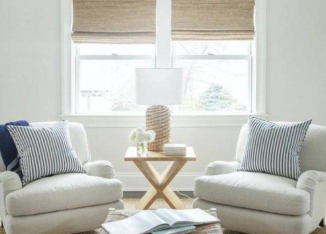 Beneath windows covered in bamboo roman shades, a rattan lamp sits on a blond x-table placed between white roll arm chairs topped with blue stripe pillows. The chairs sit on a jute rug facing a rattan stool coffee table.