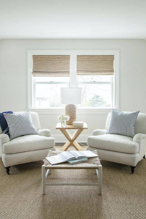 Beneath windows covered in bamboo roman shades, a rattan lamp sits on a blond x-table placed between white roll arm chairs topped with blue stripe pillows. The chairs sit on a jute rug facing a rattan stool coffee table.