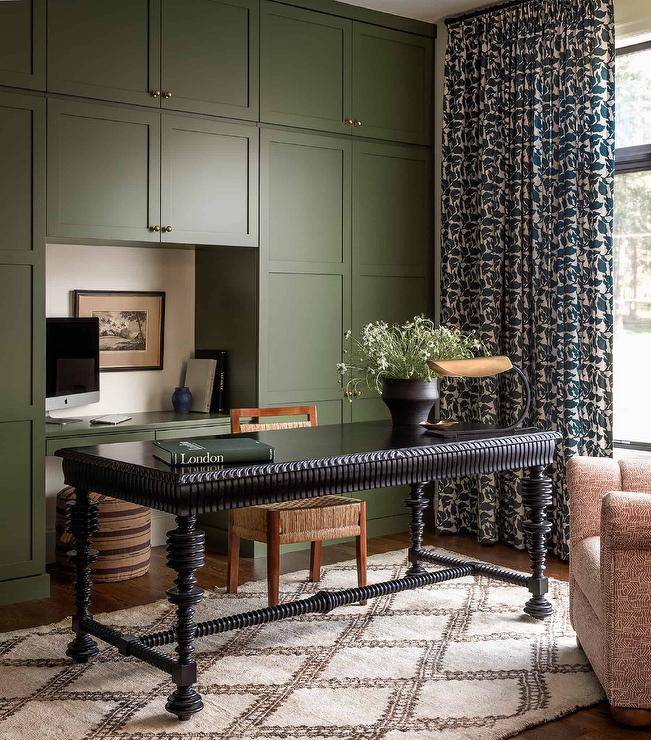 Charming home office features a black French desk placed on a trellis Moroccan rug and paired with a wood and wicker desk chair. Green built-in cabinets frame a green built-in desk paired with a tan and brown striped stool. A window is covered in white and green curtains.