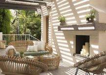 Chic long patio features a black pergola placed over modern wicker sofa and accent tables facing a pair of modern taupe outdoor chairs placed next to a white brick fireplace finished with a gray concrete mantle.