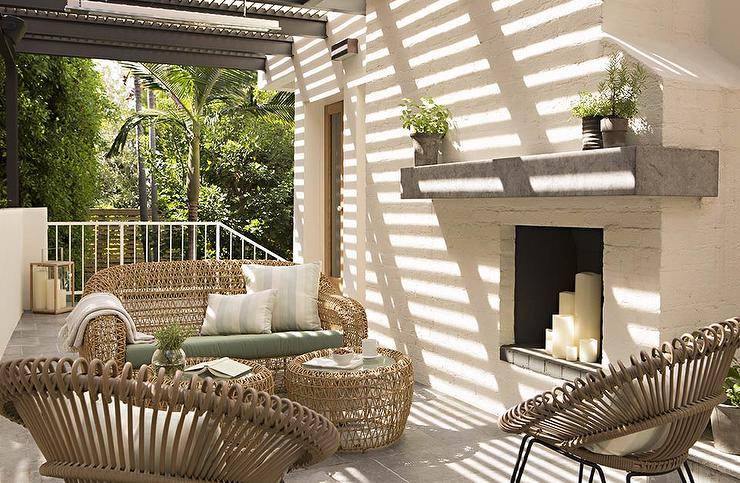 Chic long patio features a black pergola placed over modern wicker sofa and accent tables facing a pair of modern taupe outdoor chairs placed next to a white brick fireplace finished with a gray concrete mantle.