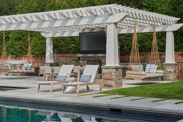 A white pergola on gray stone posts is fitted with dual hanging settees accented with gray cushions topped with white and blue pillows. In front of television mounted over a gray stone fireplace an outdoor dining table is surrounded by teak dining chairs.