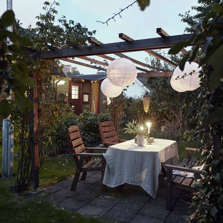 outdoor pergola at night with white hanging paper lanterns outdoor dining area with wood chairs surrounded by greenery