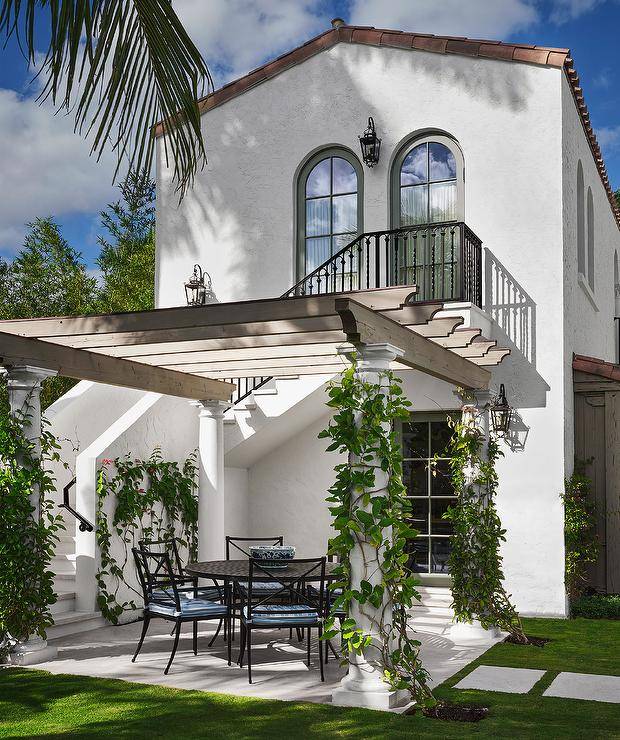 White Mediterranean style home features a pergola on Greek columns over a patio with a round wrought iron dining table and wrought iron dining chairs.