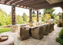 Under a pergola, iron and glass lanterns hang over a long dining table surrounded by wicker dining chairs.