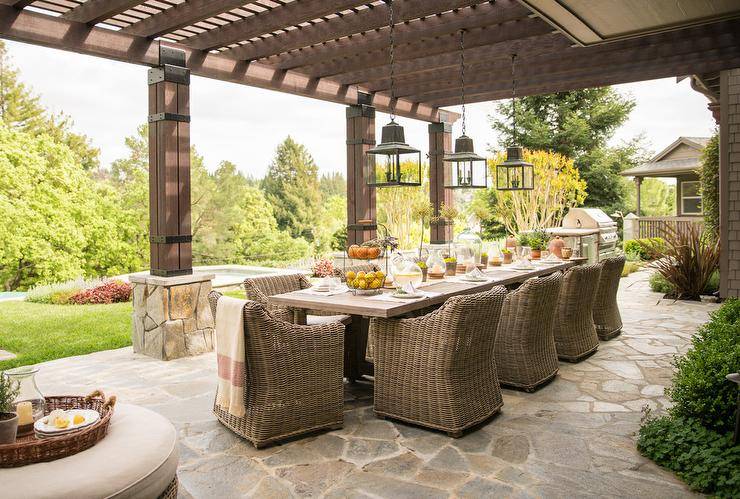 Under a pergola, iron and glass lanterns hang over a long dining table surrounded by wicker dining chairs.