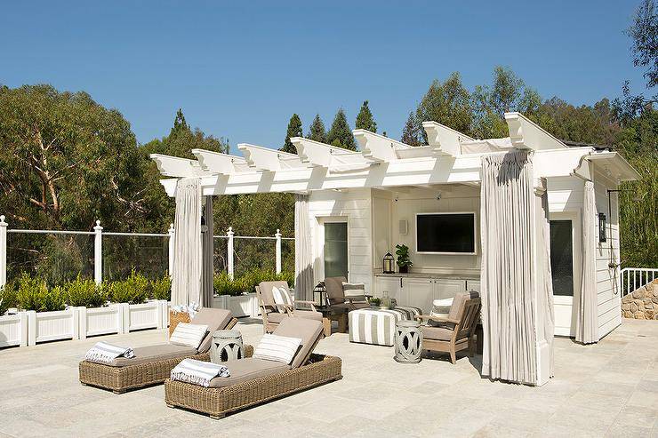 Teak loungers topped with gray cushions flank a gray rope stool placed in front of a white pool house pergola accented with gray curtains. A white and gray striped ottoman is flanked by facing side-by-side teak weathered teak chairs finished with gray cushions topped with white and gray striped pillows. A flat panel television is mounted to the side of the pool house above white built-in cabinets.