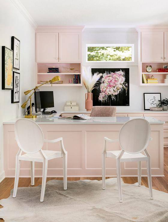 Beautifully feminine white and pink home office features white ghost chairs placed on a white cowhide rug at a pink built-in desk lit by a brass task lamp placed in front of gallery art. A black and pink art piece hangs under a window flanked by pink cabinets donning brass hardware and finished with pink shelves.