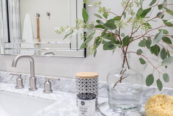 Modern nickel faucets compliment gray marble countertop and Ava vanity mirror. Countertop styling is completed with eucalyptus leaves in a glass vase, decorative candles, and a yellow sponge.