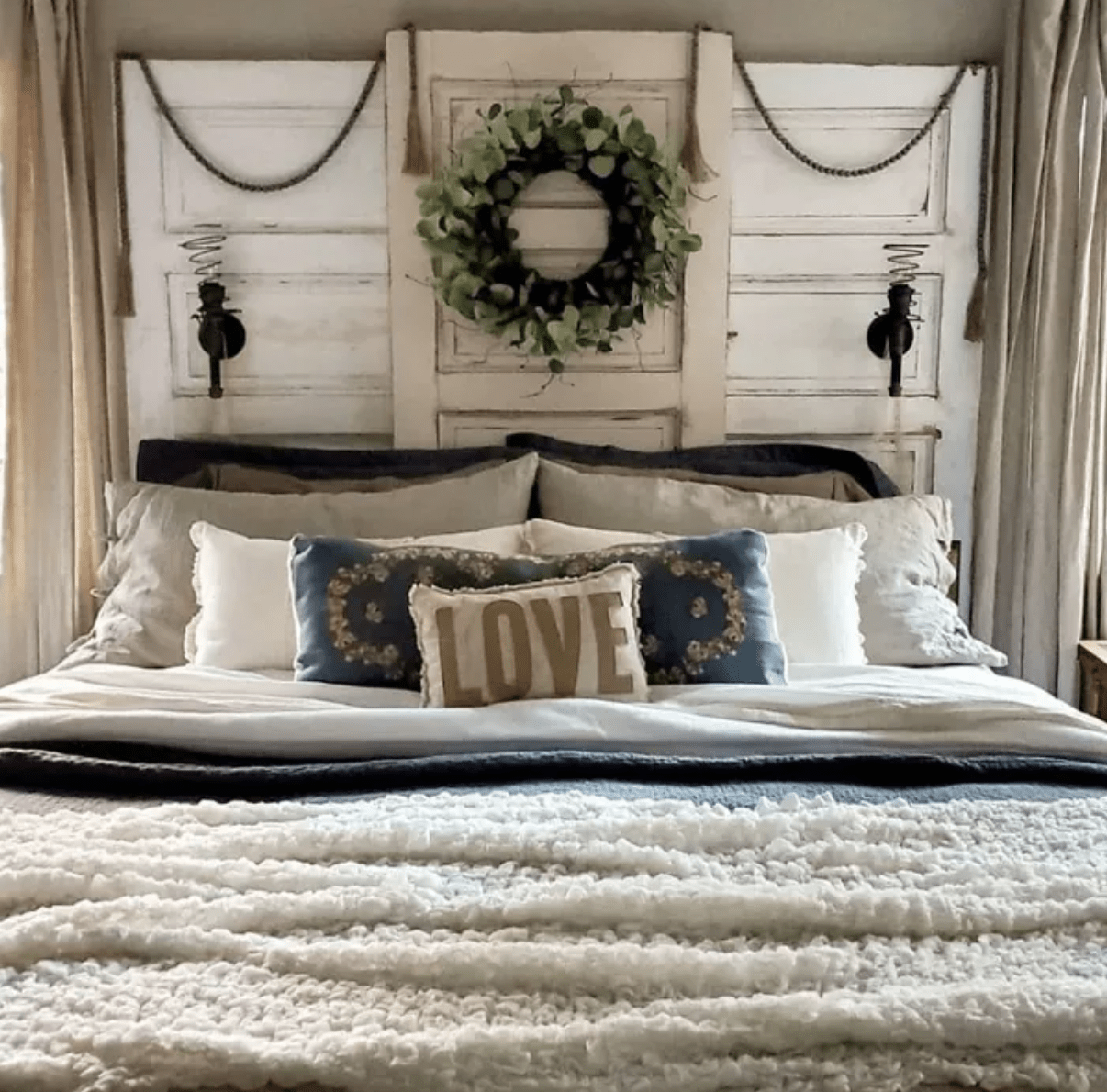 three old vintage doors are used as a bed headboard with a wreath hanging in the middle spiral wall sconces are hanging on the side love pillow fluffy fur throw