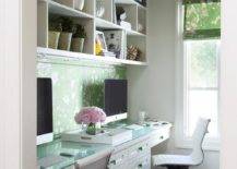 White styled shelves are mounted beneath a green painted ceiling and above a green floral backsplash located over side-by-side white built-in desk donning green glass pulls and a glass top.