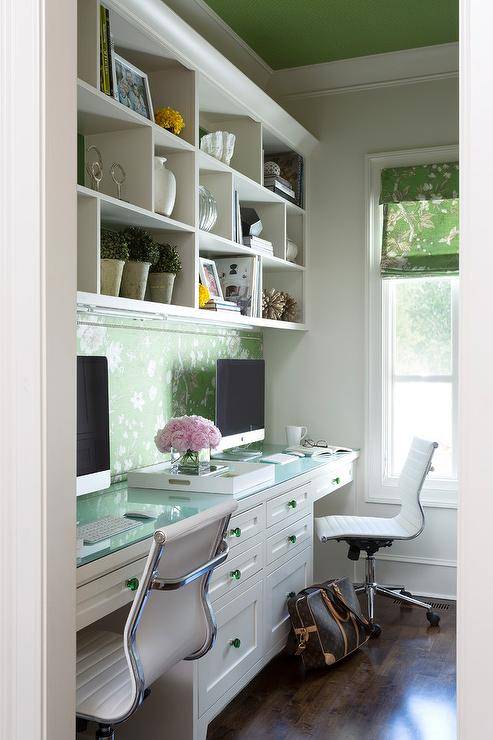 White styled shelves are mounted beneath a green painted ceiling and above a green floral backsplash located over side-by-side white built-in desk donning green glass pulls and a glass top.