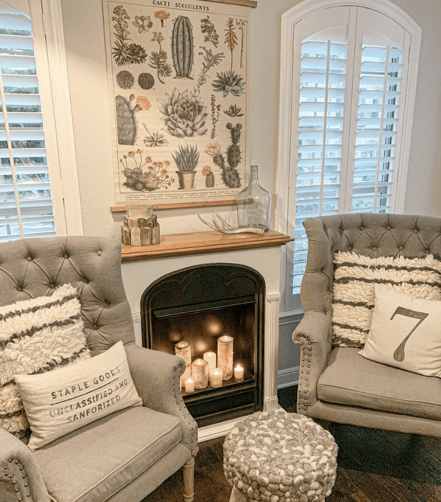 White room with small fireplace with candles inside grey tufted chairs with farmhouse pillows.