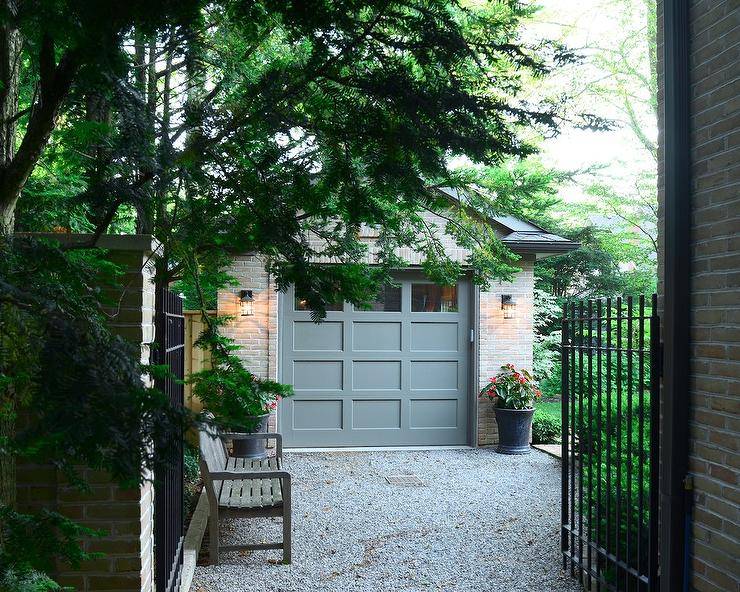 Iron gates open to a gravel driveway leading past a wooden bench to a brick garage finished with a gray multi-panel garage door flanked by carriage lanterns fixed over black planters.