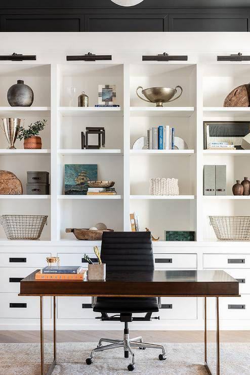 Sleek cottage black and white home office features oil rubbed bronze picture lives fixed over white built-in shelves located over white flat front drawers with oil rubbed bronze hardware. A black leather task chair sits at a stunning wood and brass desk.