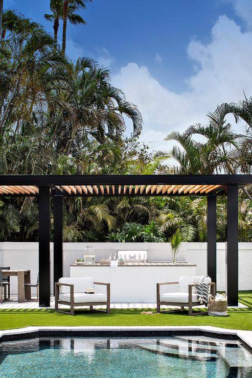 Tan and black pergola features a white outdoor kitchen and white outdoor accent chairs.
