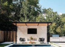 Beside an in-ground swimming pool, a white and gray rope sofa is placed beneath a black pergola with tan slats fitted over a TV niche fitted above a fireplace.