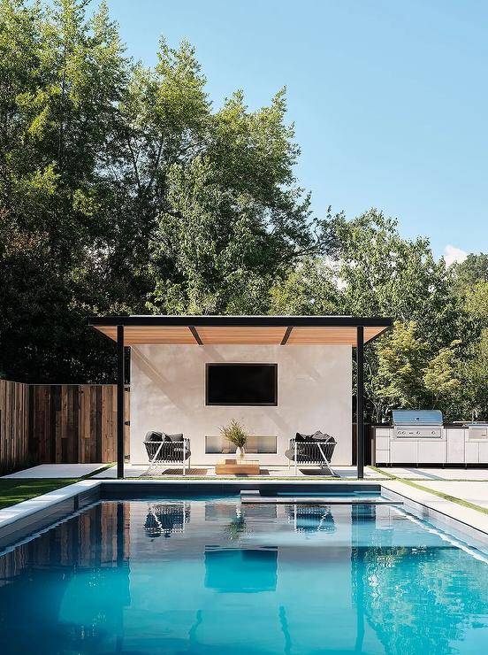 Beside an in-ground swimming pool, a white and gray rope sofa is placed beneath a black pergola with tan slats fitted over a TV niche fitted above a fireplace.