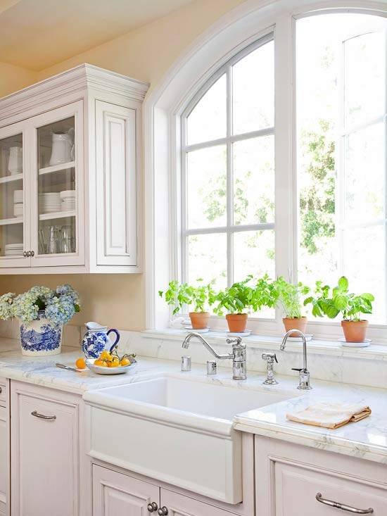 Sunny classic kitchen with pale yellow walls paint color, white kitchen cabinets with marble countertops, farmhouse sink, arched window and wood paneled dishwasher.
