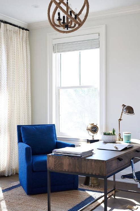 A rope sphere chandelier hangs above a wood and metal industrial desk matched with a comfy blue chair placed on a blue border jute rug in a stylish home office.