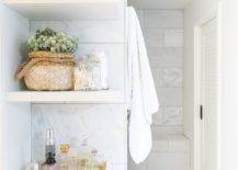 Stacked white floating bathroom shelves are mounted against staggered marble tiles over a white flat front cabinet adorned with around brass knob and topped with a perfume bottle tray.