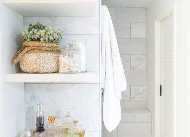 Stacked white floating bathroom shelves are mounted against staggered marble tiles over a white flat front cabinet adorned with around brass knob and topped with a perfume bottle tray.
