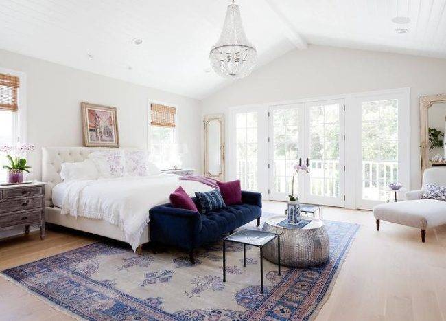 Elegant bedroom features a white beaded glass chandelier hung from a vaulted shiplap ceiling over a blue velvet tufted sofa topped with purple velvet accent pillows positioned in front of a white tufted bed dressed in a white ruffled bedding. A painting hangs above the bed flanked by windows covered in bamboo roman shades positioned above long wood and marble dressers while a pink and blue Turkish rug sits under the bed covering wide plank blond wood floors and also below a hammered metal coffee table located between black leather stools. Beside a reading nook, the room is completed with distressed French floor mirrors are located on either side of glass paneled French patio door.