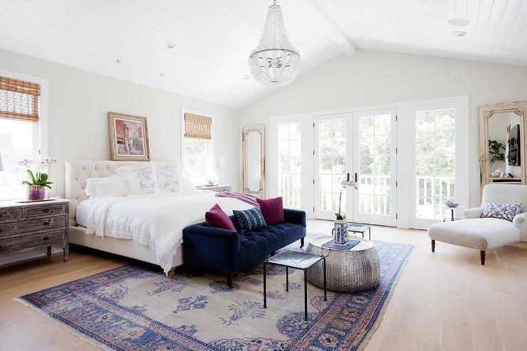 Elegant bedroom features a white beaded glass chandelier hung from a vaulted shiplap ceiling over a blue velvet tufted sofa topped with purple velvet accent pillows positioned in front of a white tufted bed dressed in a white ruffled bedding. A painting hangs above the bed flanked by windows covered in bamboo roman shades positioned above long wood and marble dressers while a pink and blue Turkish rug sits under the bed covering wide plank blond wood floors and also below a hammered metal coffee table located between black leather stools. Beside a reading nook, the room is completed with distressed French floor mirrors are located on either side of glass paneled French patio door.