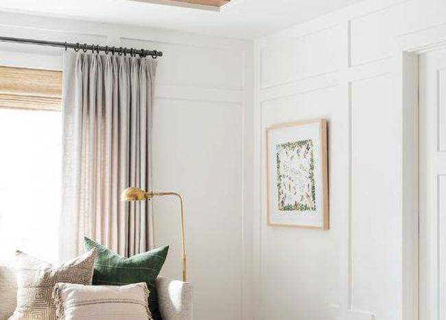 Gray curtains are layered in front of a bamboo roman shade hung above an oatmeal linen sofa lit by a brass pharmacy lamp. A black stripe cube ottoman sits beside the sofa, in front of a board and batten wall, and beneath a ceiling finished with stained wood beams.