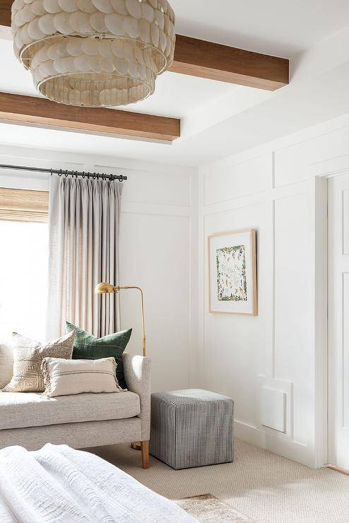 Gray curtains are layered in front of a bamboo roman shade hung above an oatmeal linen sofa lit by a brass pharmacy lamp. A black stripe cube ottoman sits beside the sofa, in front of a board and batten wall, and beneath a ceiling finished with stained wood beams.