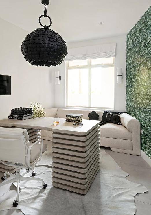Chic modern home office light gray sofa placed beneath a window covered in a white roman shade and facing a modern gray desk lit by a black discs chandelier. A white task chair sits at the desk on a white cowhide rug, while an accent wall is covered in green stalking tiger wallpaper.
