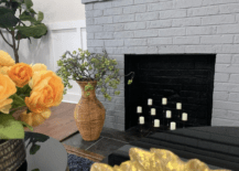 white candles in grey brick fireplace with wicker vase and greenery