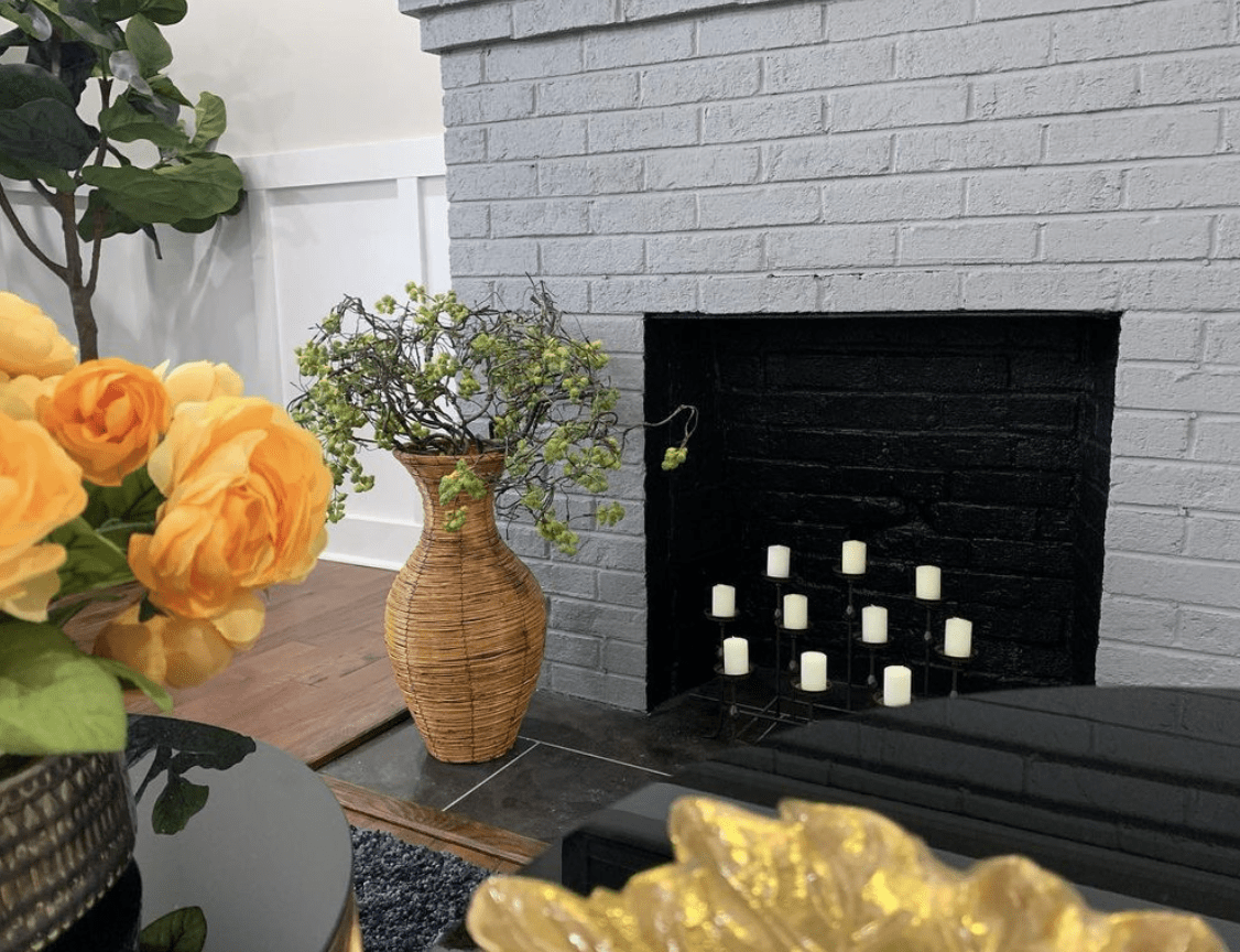 white candles in grey brick fireplace with wicker vase and greenery