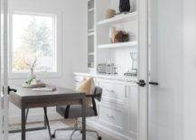 Glass French doors open to a home office boasting a gray desk chair placed on a gray suede rug at a wood and metal desk positioned in front of white built-in cabinets fixed beneath shelves flanked by glass front cabinets.