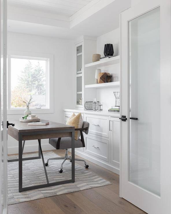 Glass French doors open to a home office boasting a gray desk chair placed on a gray suede rug at a wood and metal desk positioned in front of white built-in cabinets fixed beneath shelves flanked by glass front cabinets.