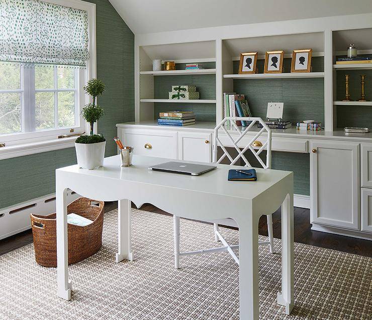 White and green home office features built in shelves on green grasscloth wallpaper, a white baroque desk and a white bamboo desk chair over a tan rug.