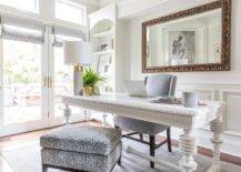 Beautifully designed home office features a white spindle desk placed on a gray rug and matched with a gray wingback chair and a leopard print pillow top bench. Behind the desk, a gilt French mirror hangs from a white wall lined with wainscoting. White shelves are built-in at the end of the wall.