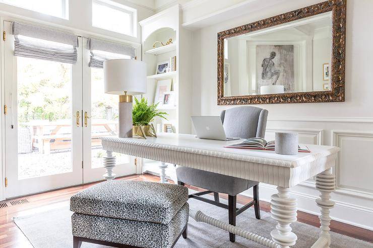 Beautifully designed home office features a white spindle desk placed on a gray rug and matched with a gray wingback chair and a leopard print pillow top bench. Behind the desk, a gilt French mirror hangs from a white wall lined with wainscoting. White shelves are built-in at the end of the wall.