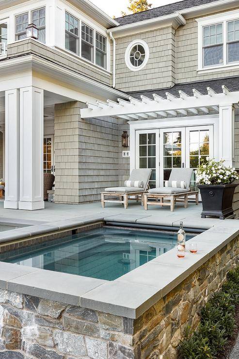 Rustic stone clad spa is accented with gray pool line tiles and positioned in front of two pool loungers with gray cushions topped with gray striped bolster pillows. The loungers sit beneath a white pergola.