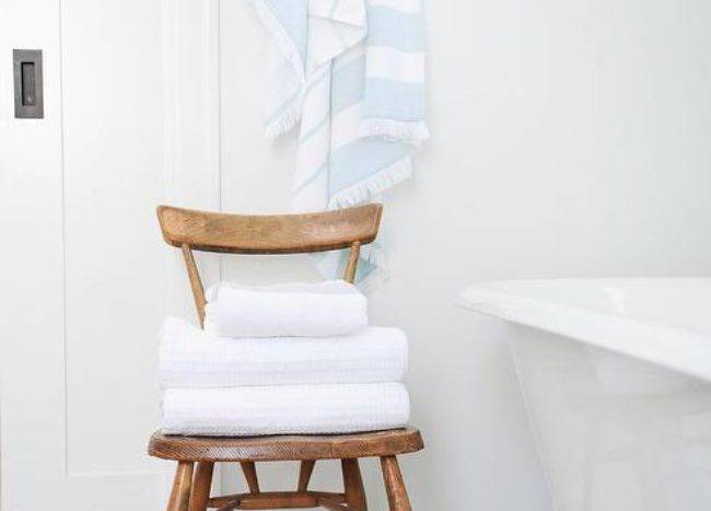 White and gray bathroom styled with a brown wooden vintage chair storing folded towels beside a white clawfoot tub atop large gray hexagon floor tiles.