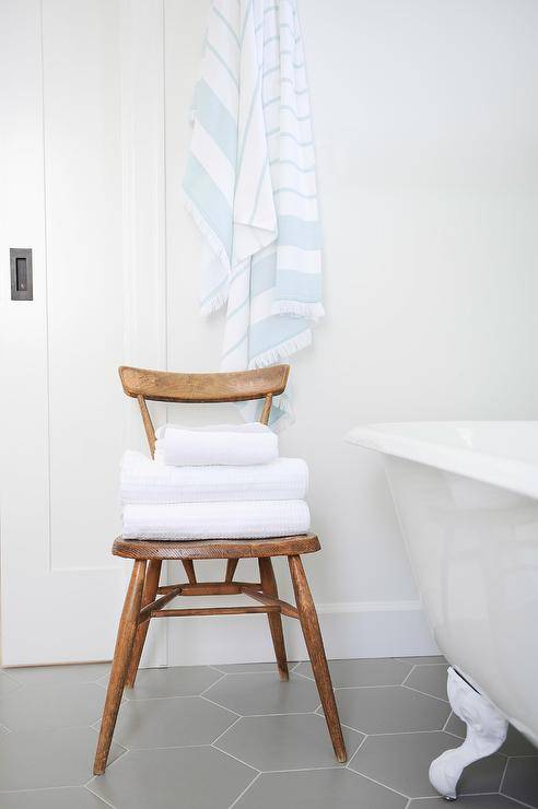 White and gray bathroom styled with a brown wooden vintage chair storing folded towels beside a white clawfoot tub atop large gray hexagon floor tiles.