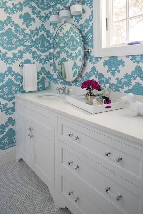 Gorgeous white and turquoise bathroom with turquoise flocked wallpaper framing a nickel vanity light over a round pivot wall mirror above the shaker front vanity with white counters and glass drawer pulls atop white penny tiled floors.