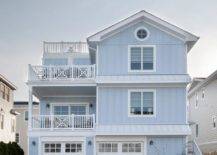 Periwinkle beach house boasting white trim with two white x-trimmed balconies, vertical siding, white double garage doors and gray pavers.