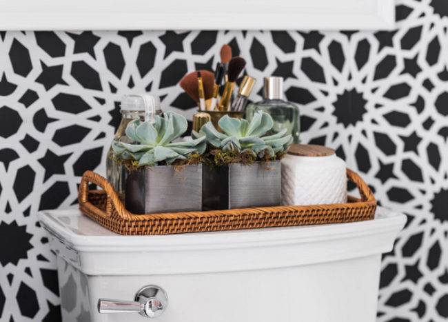 Black and white bathroom features an accent wall clad in Schumacher Agadir Screen Noir Wallpaper lined with a toilet topped with a woven tray filled with succulents.