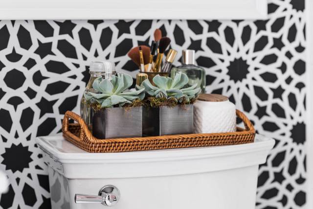 Black and white bathroom features an accent wall clad in Schumacher Agadir Screen Noir Wallpaper lined with a toilet topped with a woven tray filled with succulents.